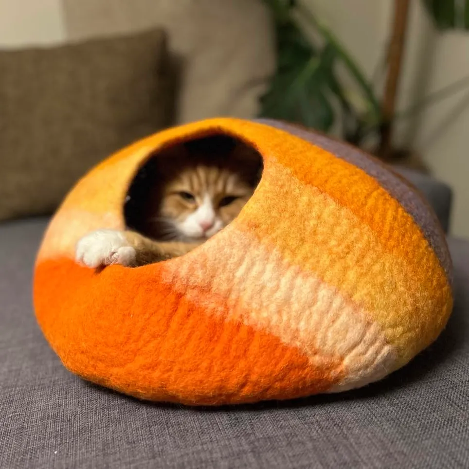 Regnbuen kattehule i oransje, gul og kremhvit tovet ull, med en rødhvit katt inne i hulen